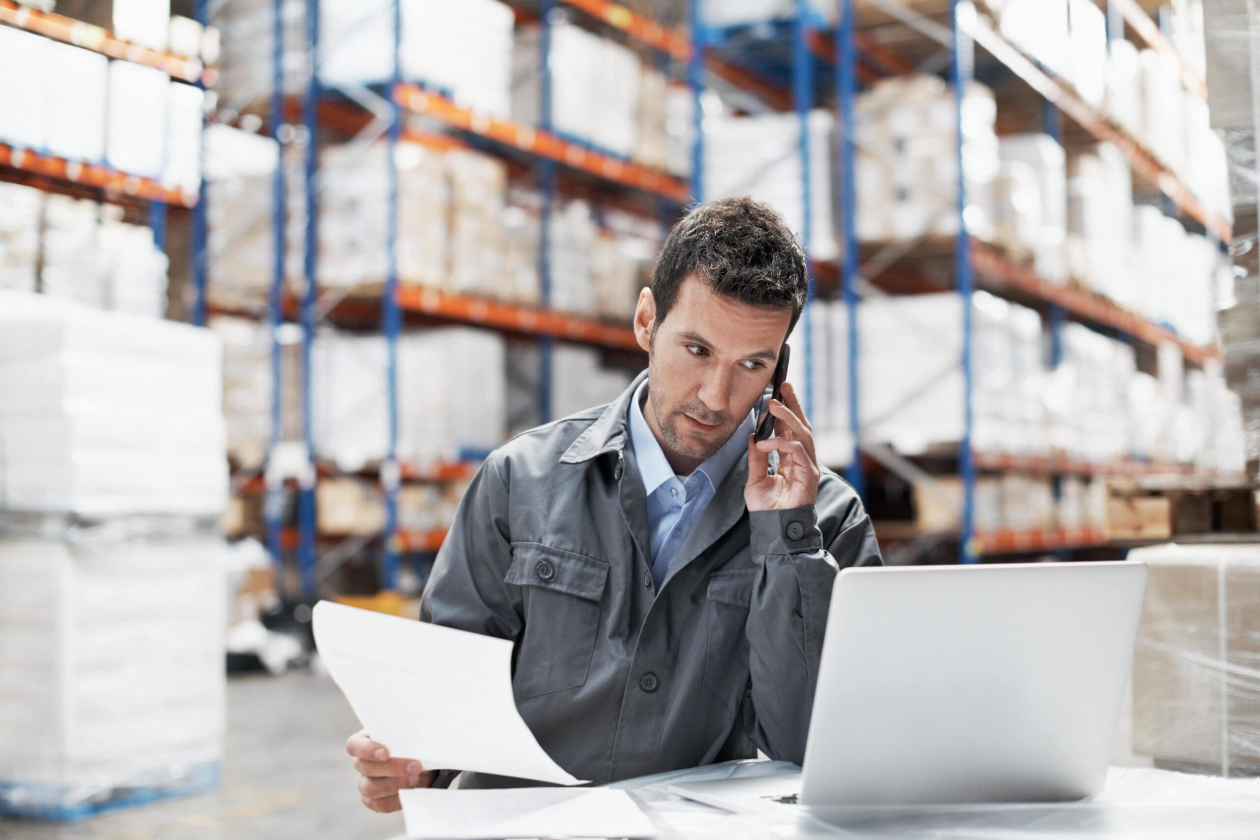 Photo of a man in a warehouse talking on a phone while looking at some papers and a laptop.
